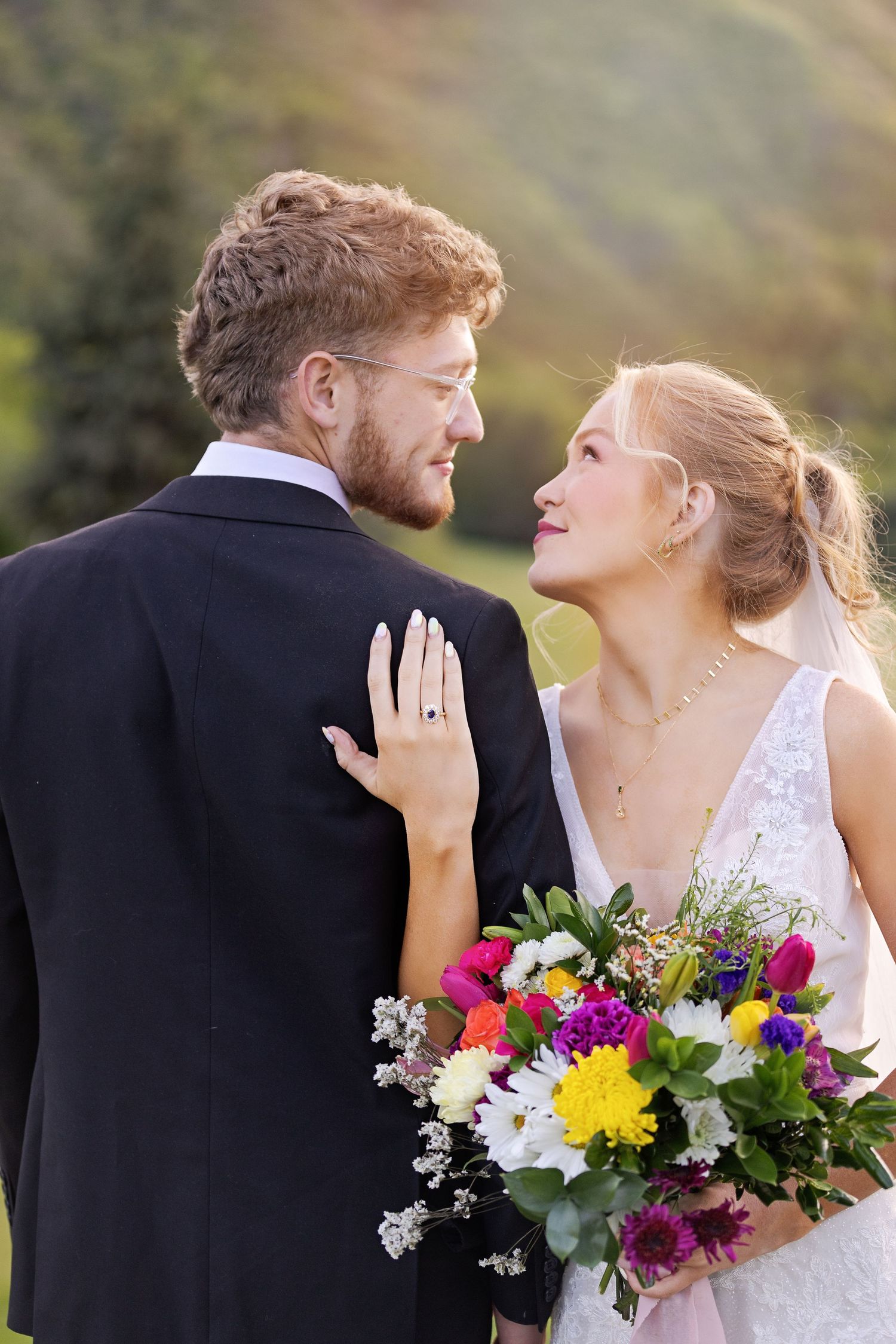 Newlywed couple in wedding attire embrace outdoors while bride holds colorful wildflower bouquet against scenic backdrop.