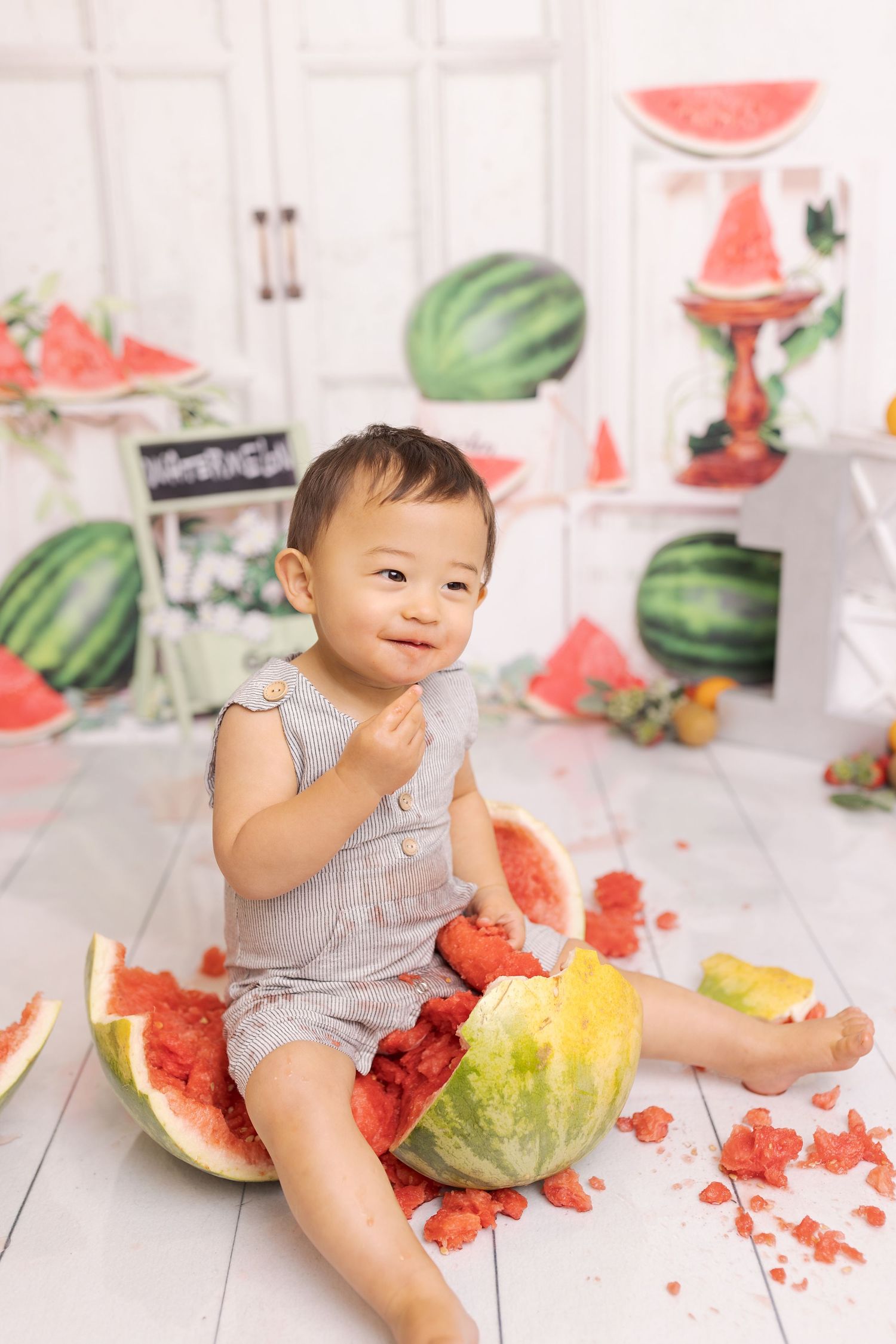 Sweet child in light gray romper sitting with fresh watermelon slices in a summery white studio setting.