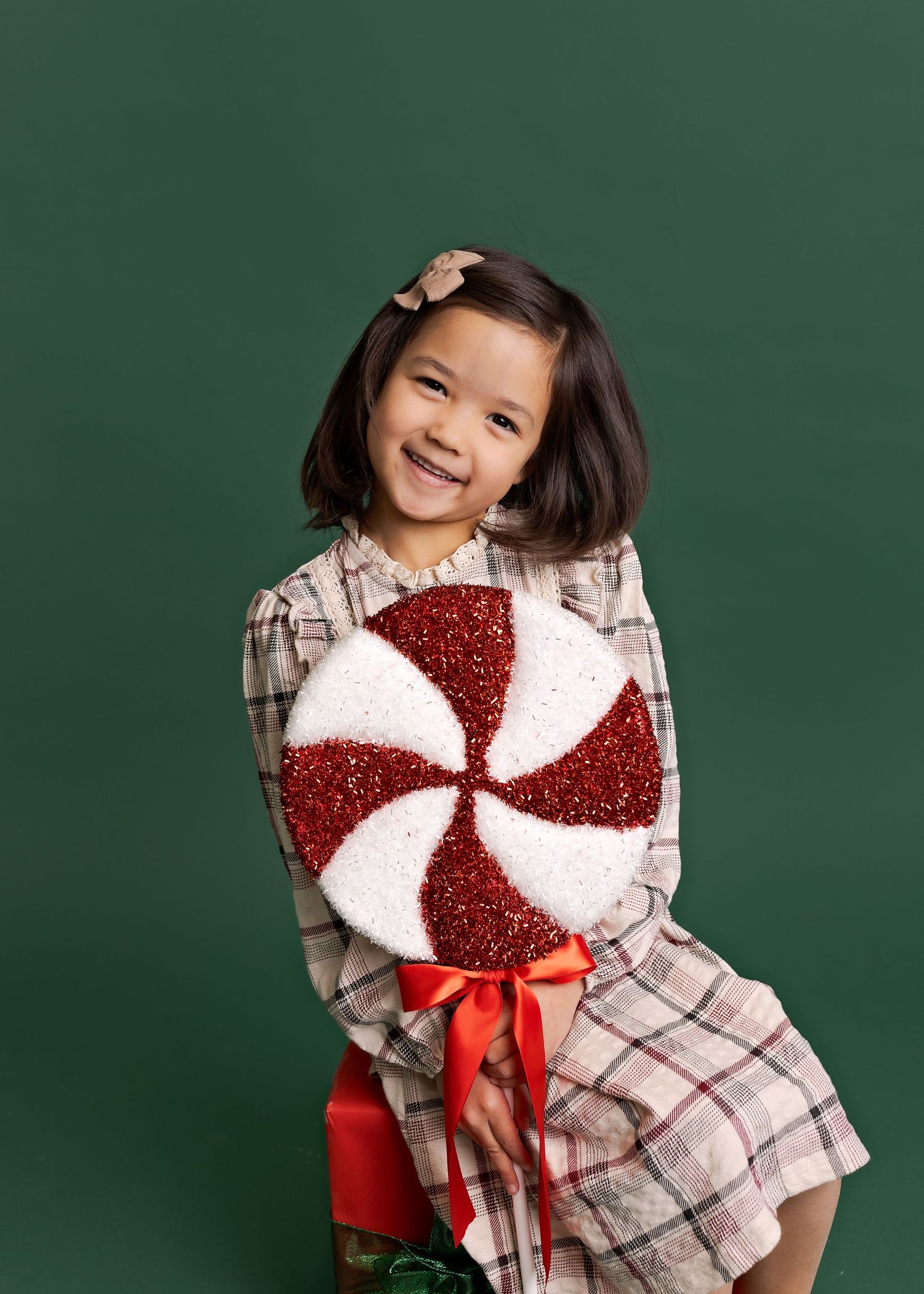 A child wearing a plaid dress and sparkly red peppermint bow sits against a green backdrop in a holiday photo session.