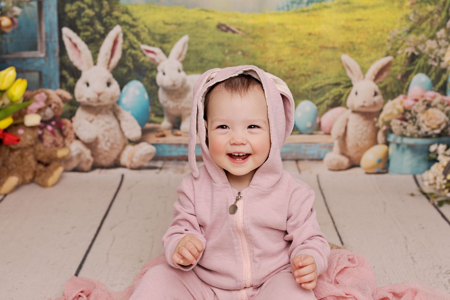 Adorable baby in pink outfit sits on wooden deck surrounded by plush Easter bunny decorations and spring scenery backdrop.