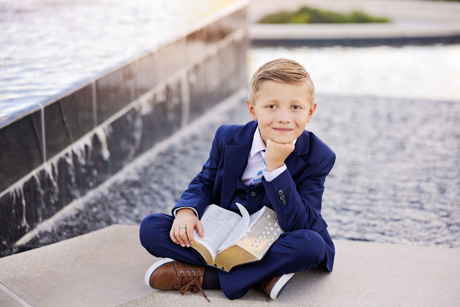 A child in a navy suit sits cross-legged with an open book outdoors.