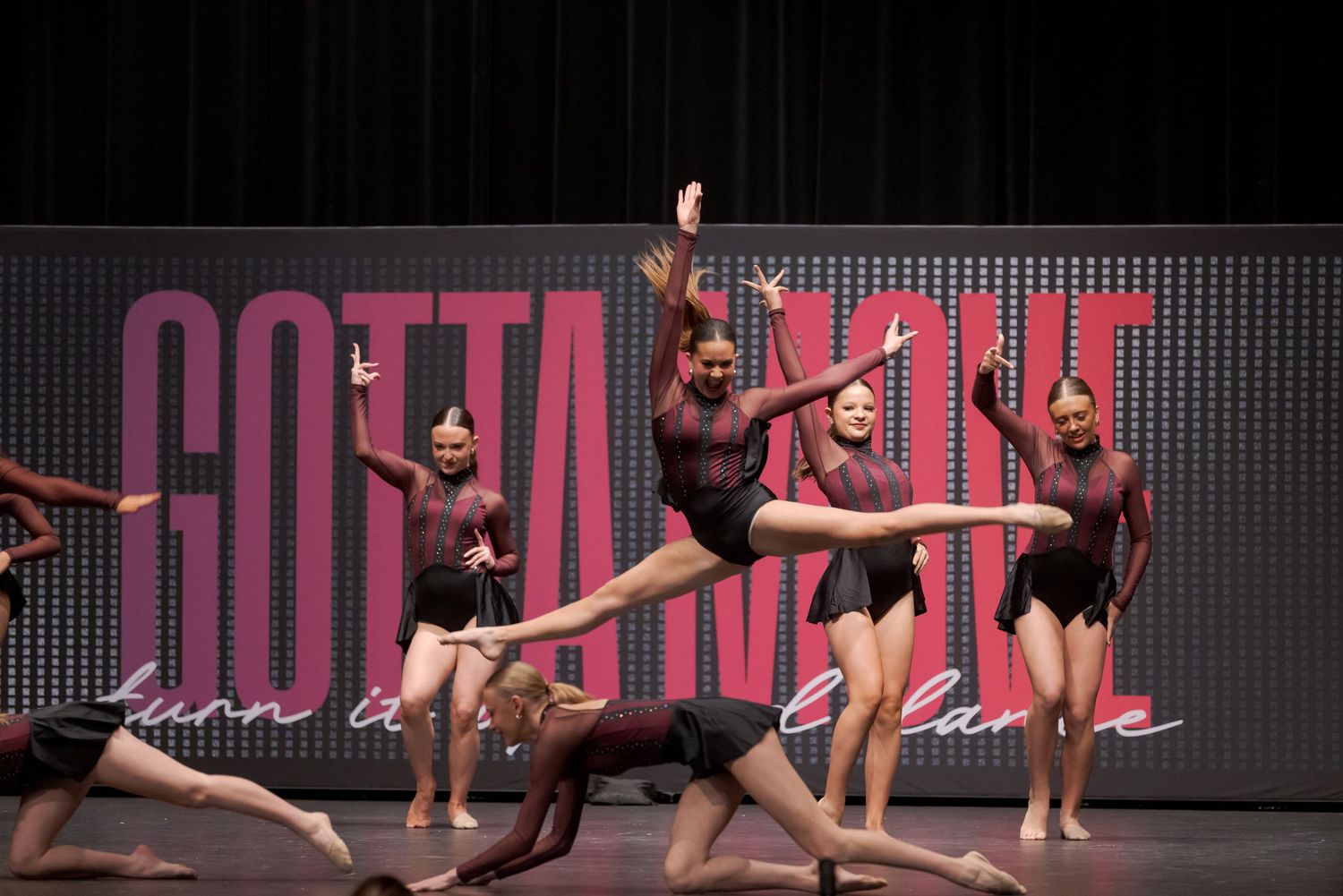 Athletic dancers perform a synchronized contemporary dance routine on a dark stage with dramatic red lighting.