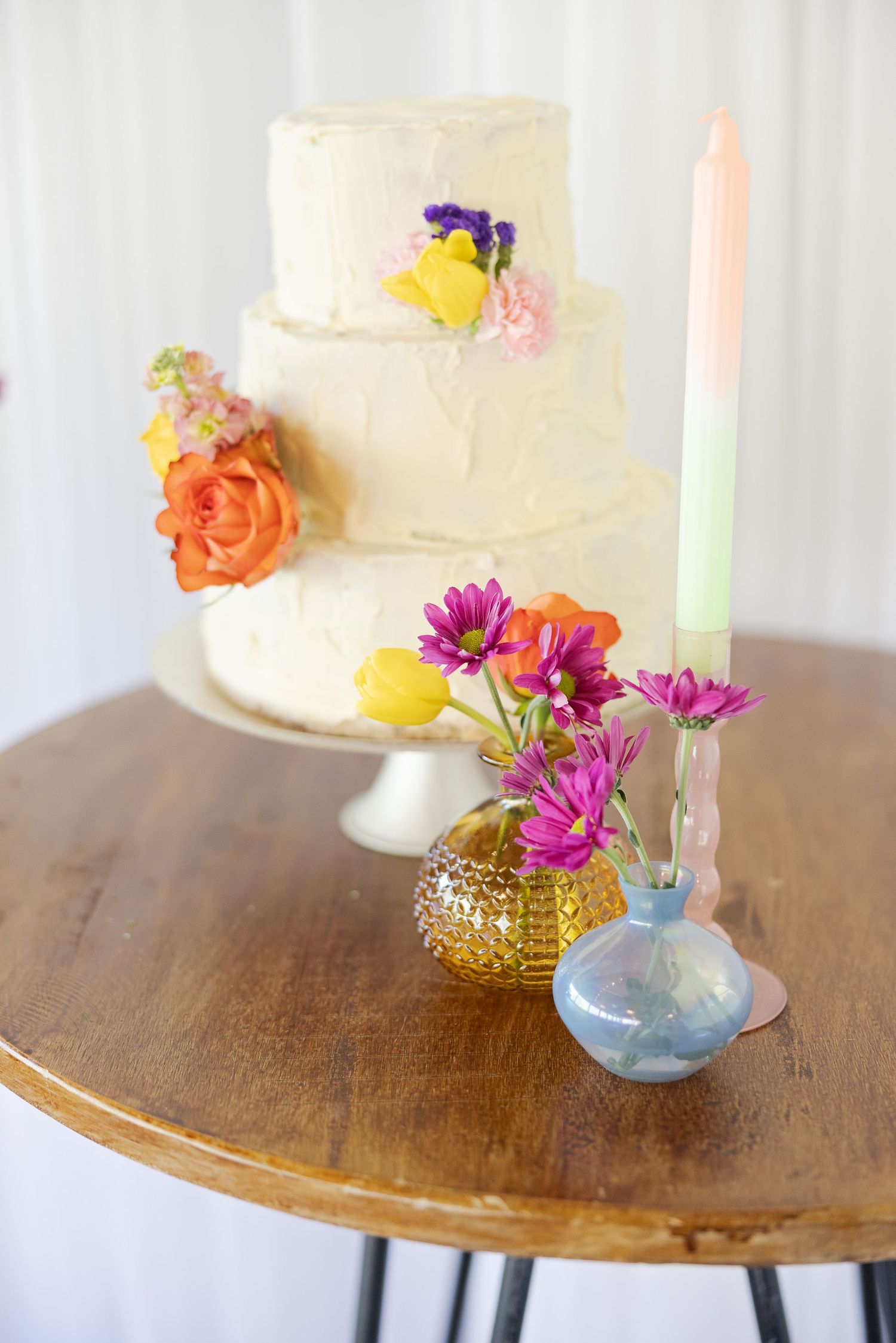 A round wooden table displays a white frosted cake with vibrant orange roses and pink daisies in glass bud vases.
