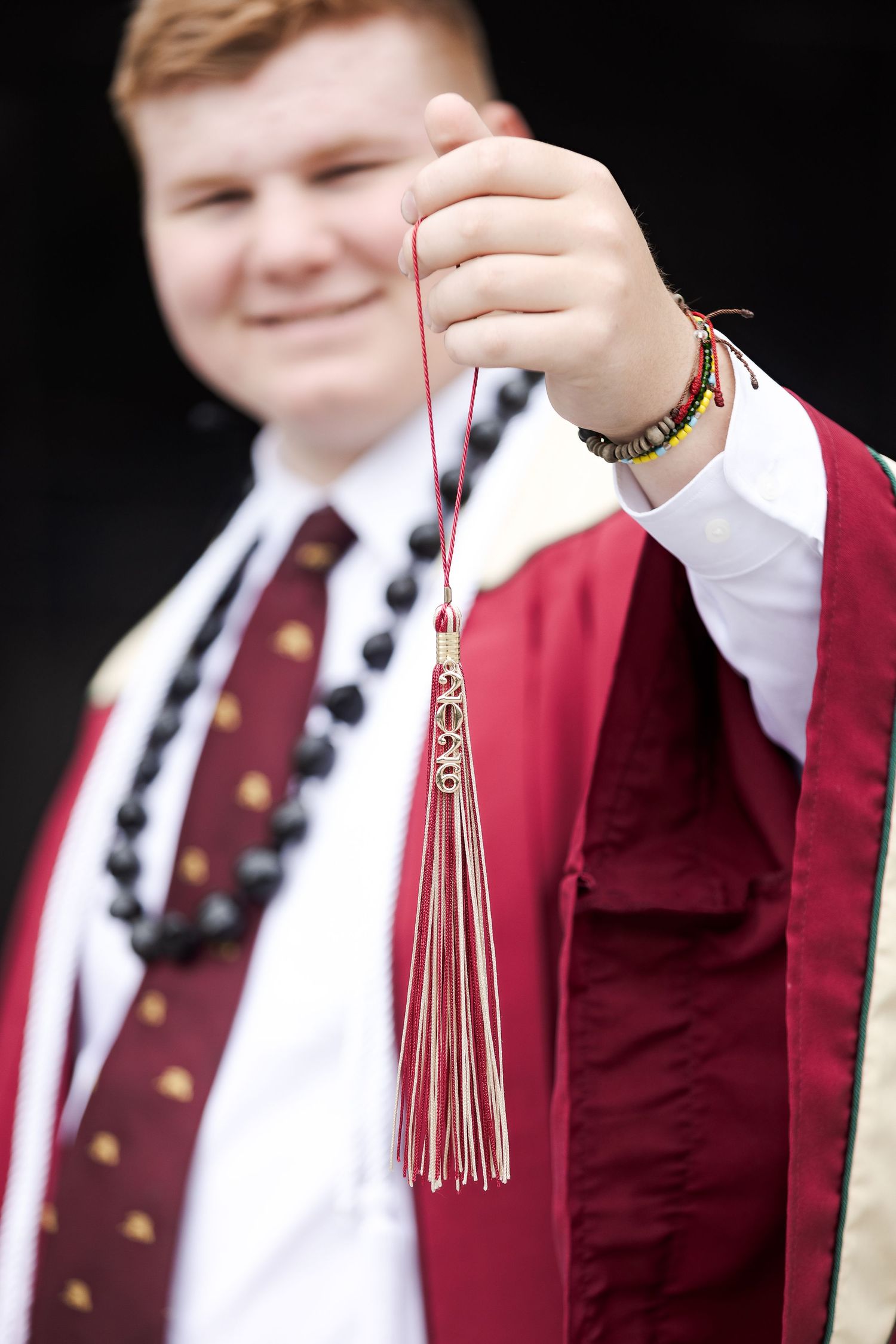 Smiling graduate in maroon gown holds tassel, wearing beaded necklace and bracelets, celebrating academic achievement.