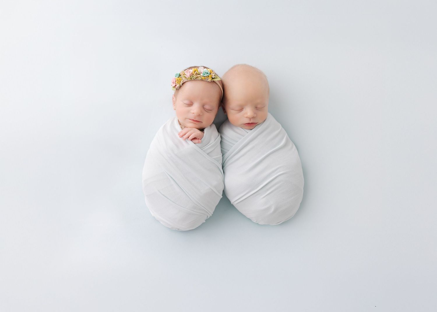 Two swaddled newborn babies laying side by side on white background with one wearing a small floral crown.