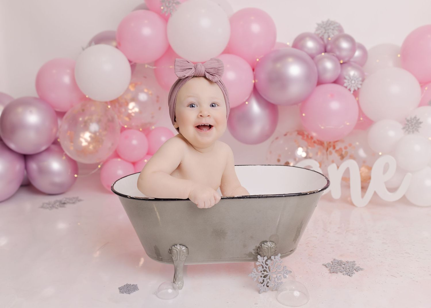 Baby in vintage bathtub with pink and silver balloon backdrop during milestone photoshoot.