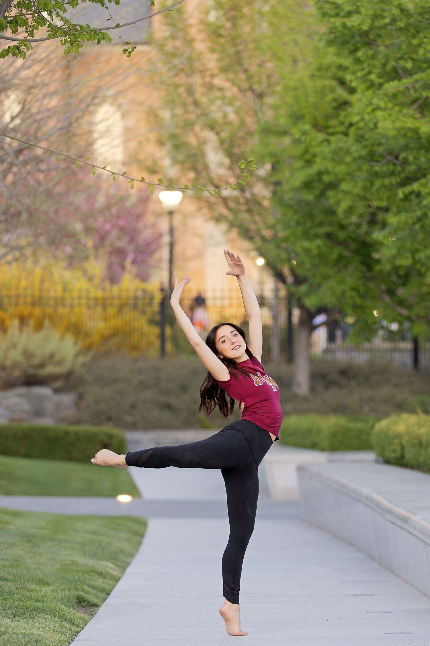 Graceful dancer practices high kicks and dance moves outdoors on stone path surrounded by trees and foliage.