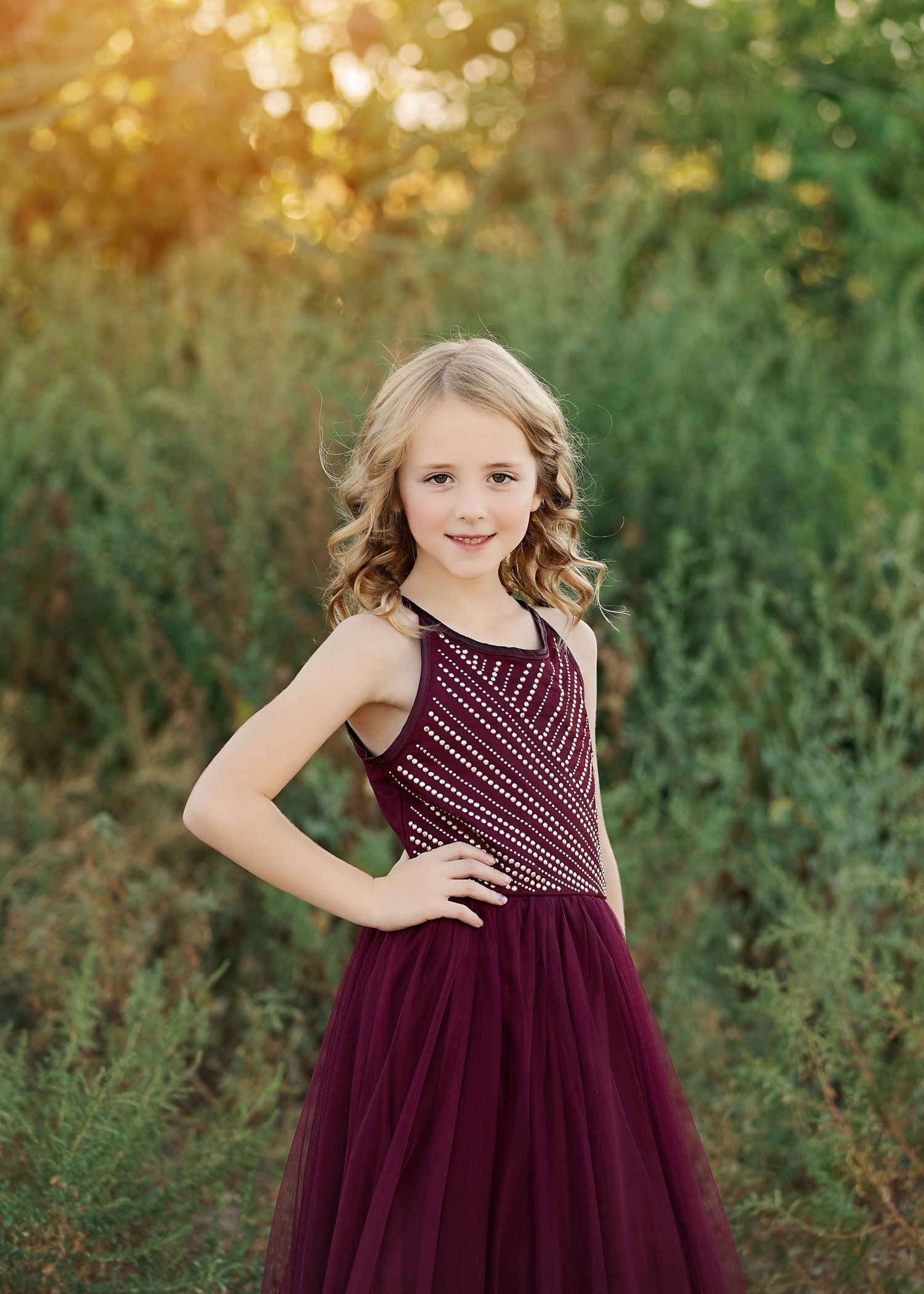 Young person in burgundy formal dress poses against blurred natural background at sunset.