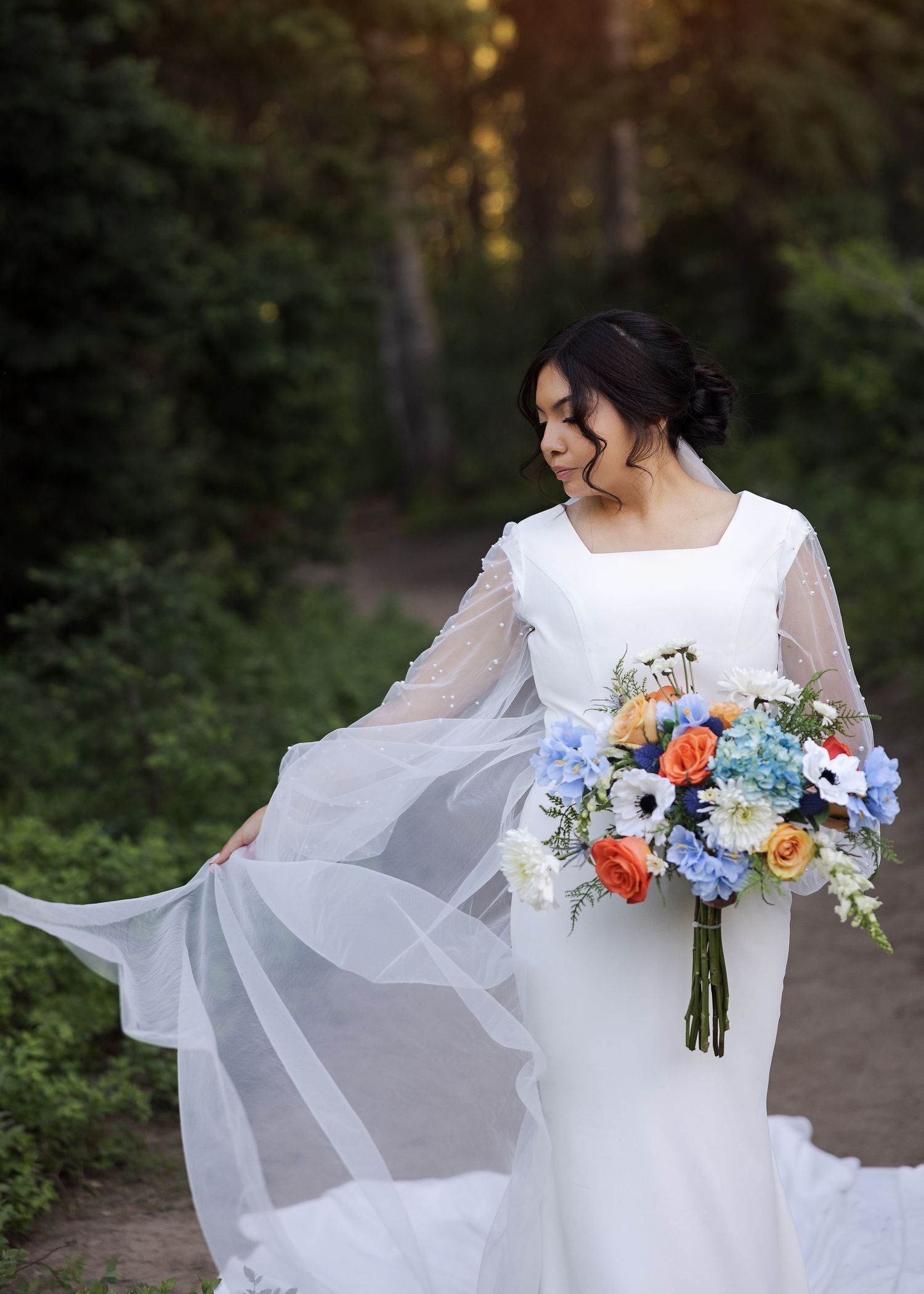 Elegant white wedding dress with sheer sleeves flows in the forest as the bride holds a vibrant wildflower bouquet.
