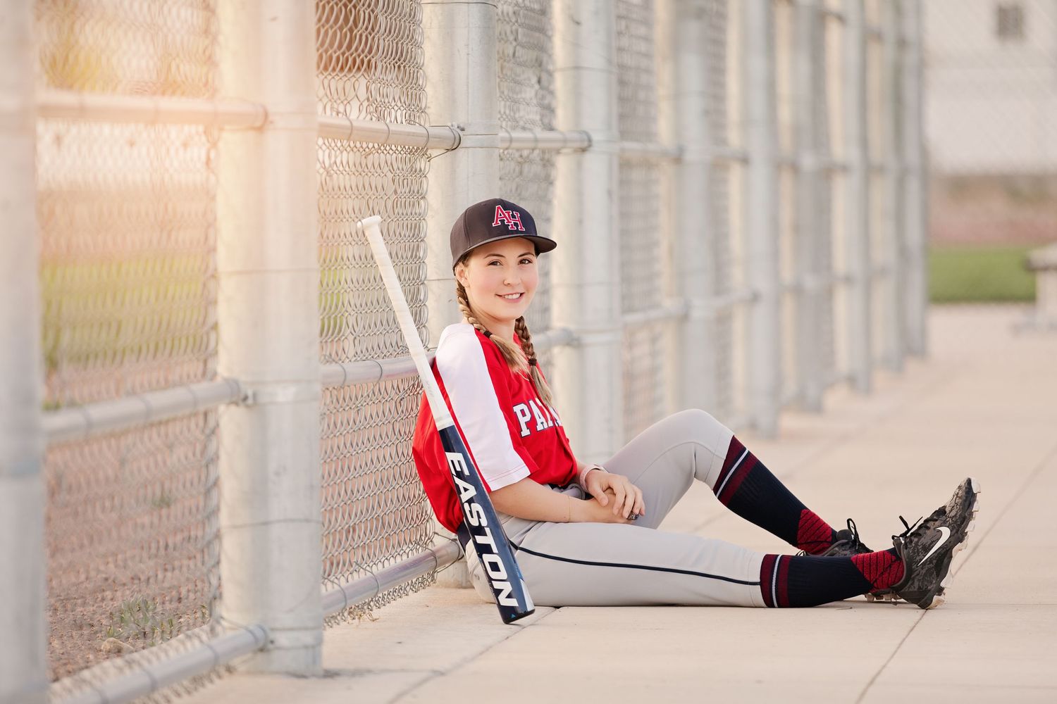 Softball player in red uniform sitting against chain link fence.