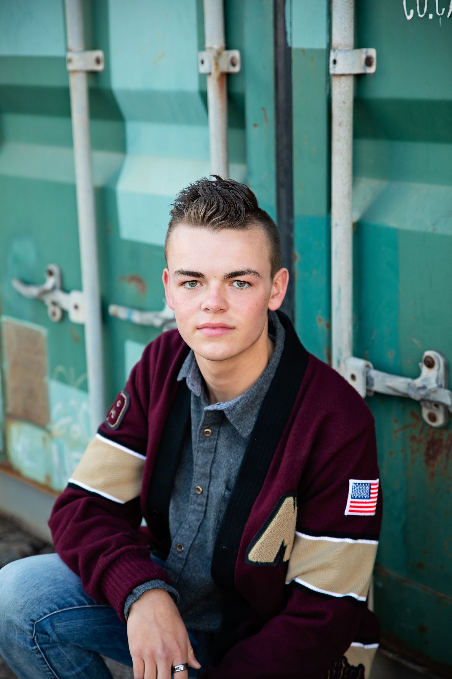 Portrait against turquoise container wearing maroon letterman jacket and denim shirt.