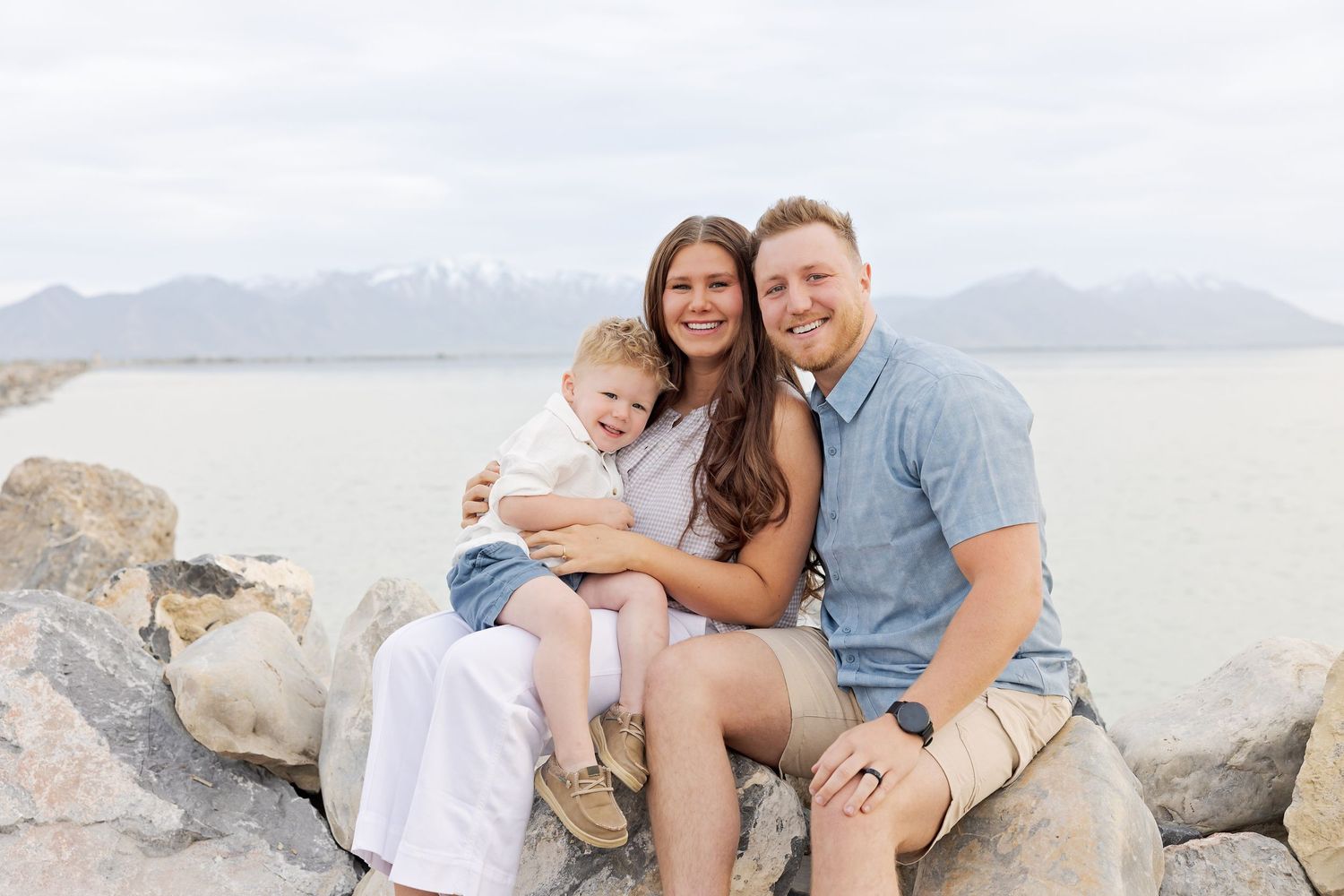 Happy family of three sitting on rocks by a lake with mountain views, smiling couple holding young toddler outdoors.