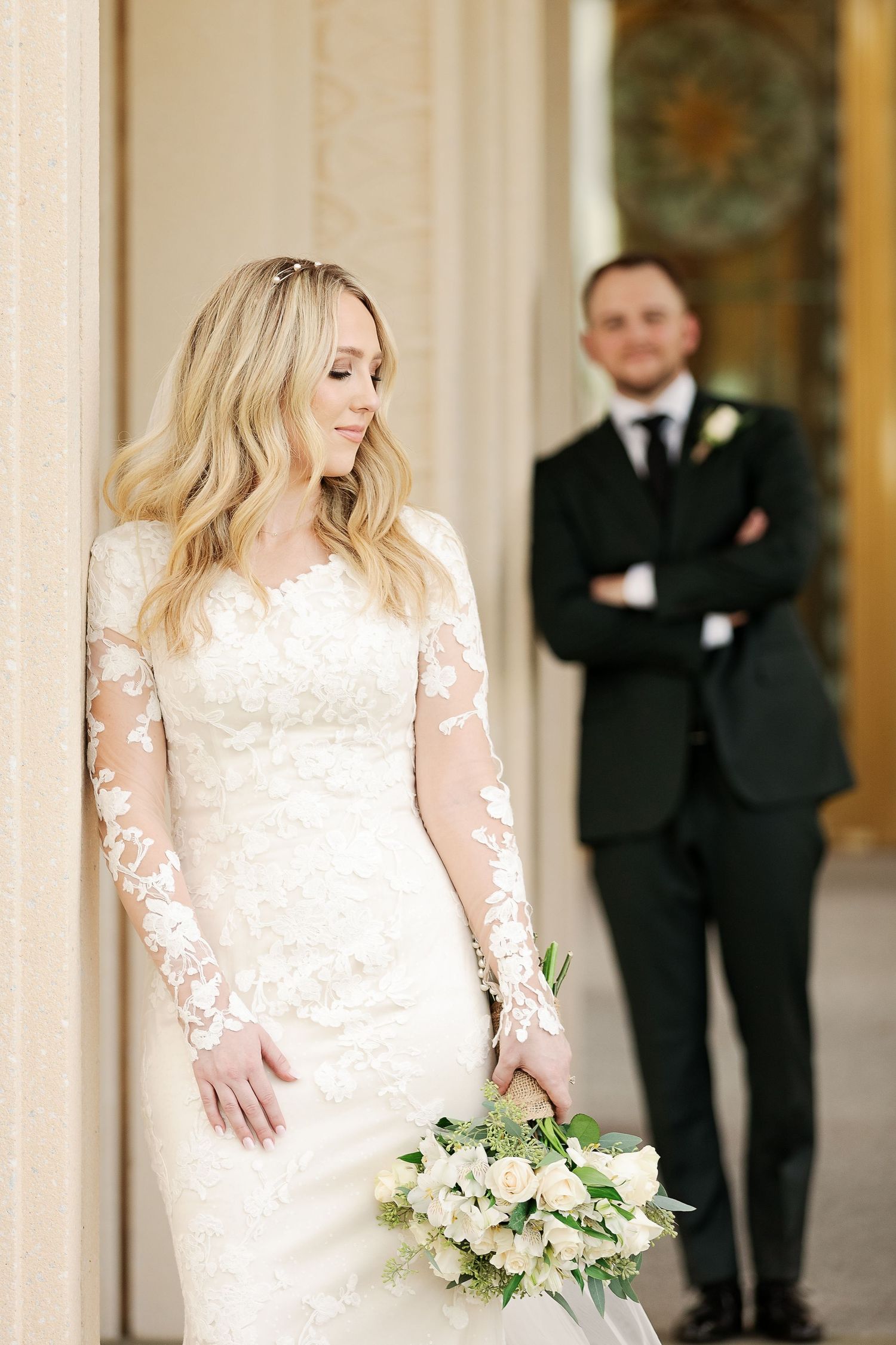 Bride in lace gown holds bouquet while groom stands in background.