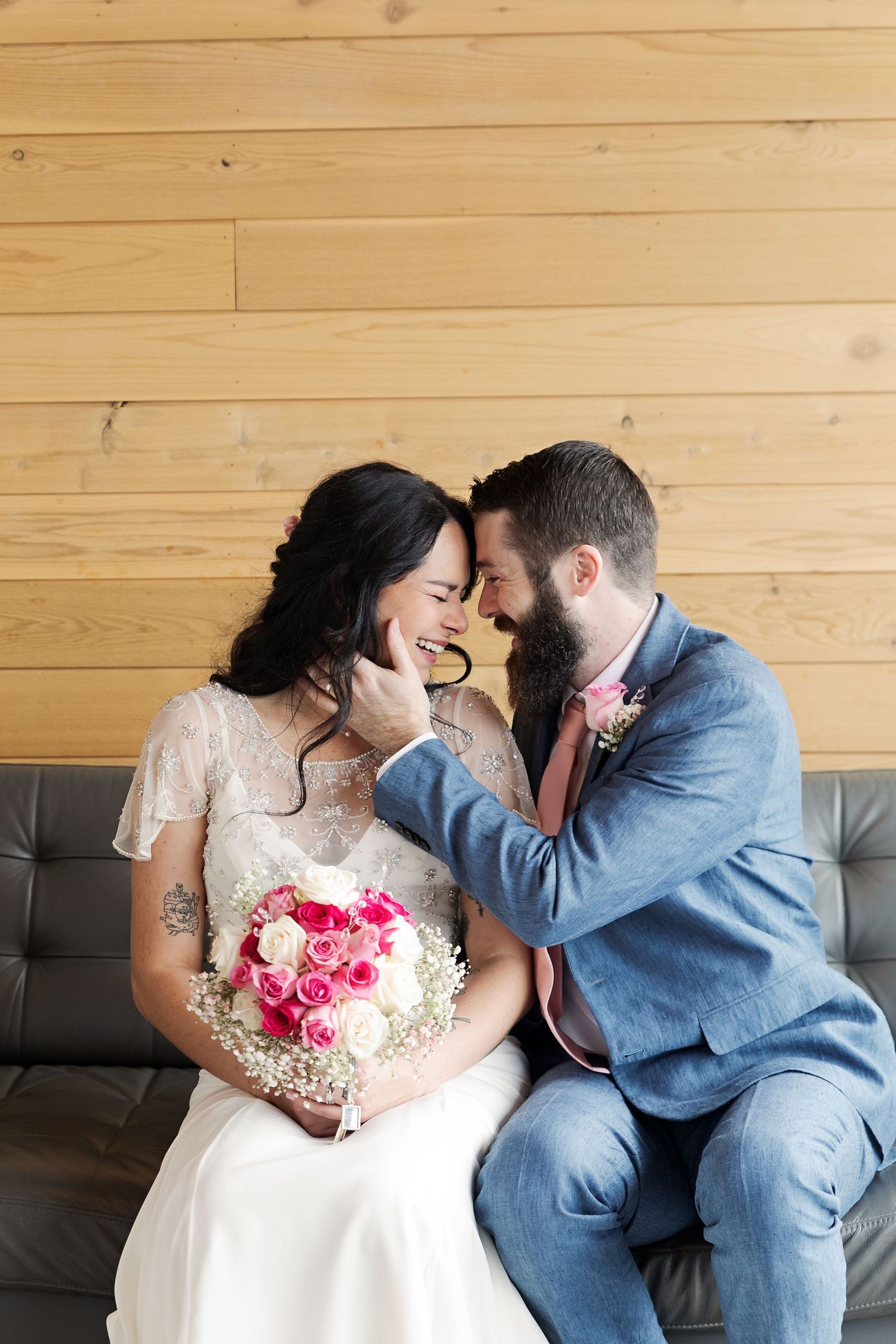 Intimate wedding portrait on grey couch against wooden wall with pink and white bouquet.