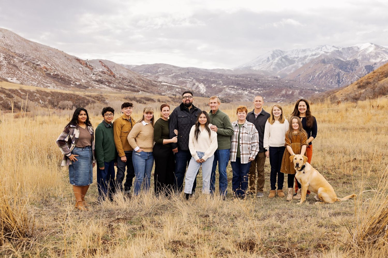 A group stands together in a scenic mountain field with golden grass and snow-capped peaks in the background.