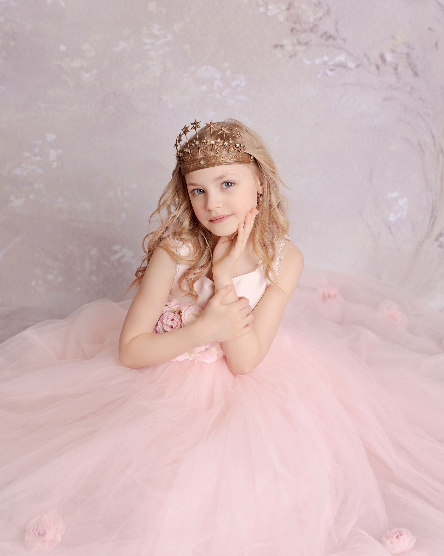 A young person in a pink tulle ball gown and tiara poses against a textured gray backdrop.