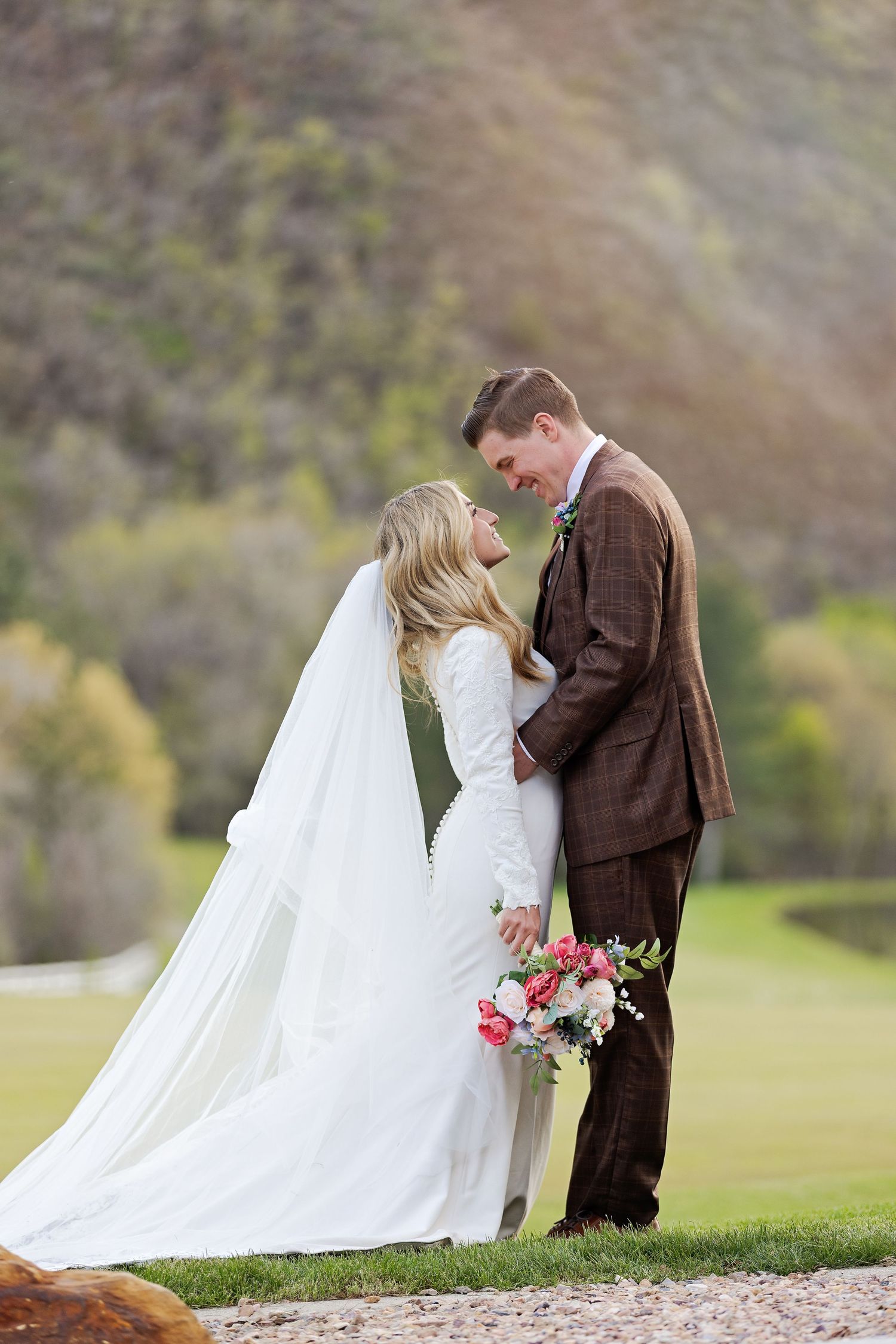 A romantic wedding portrait shows a couple in white dress and brown suit holding flowers on a grassy hill in nature.