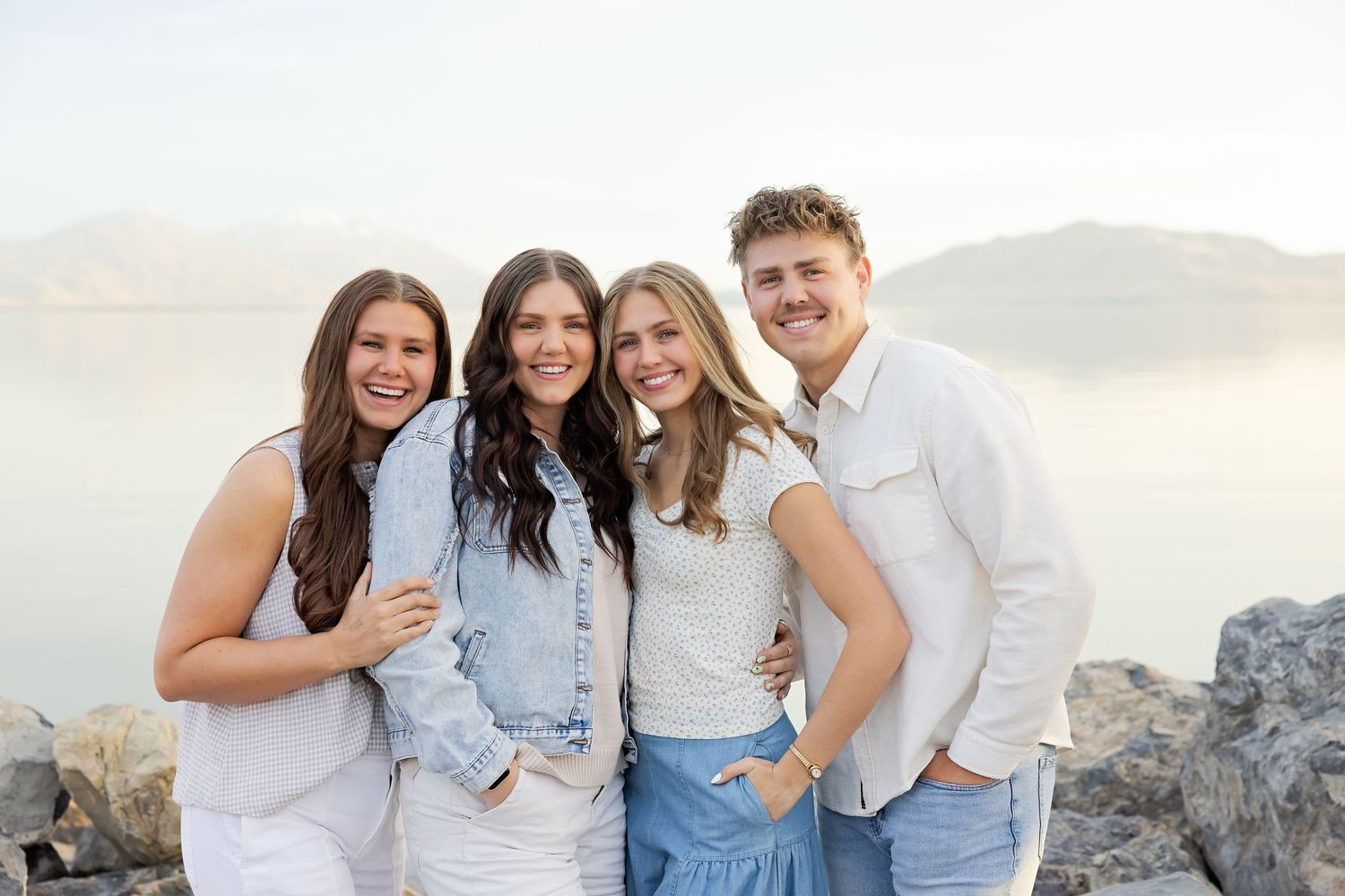 Four smiling young adults pose together outdoors near a rocky shore with misty mountains and calm water in the background.