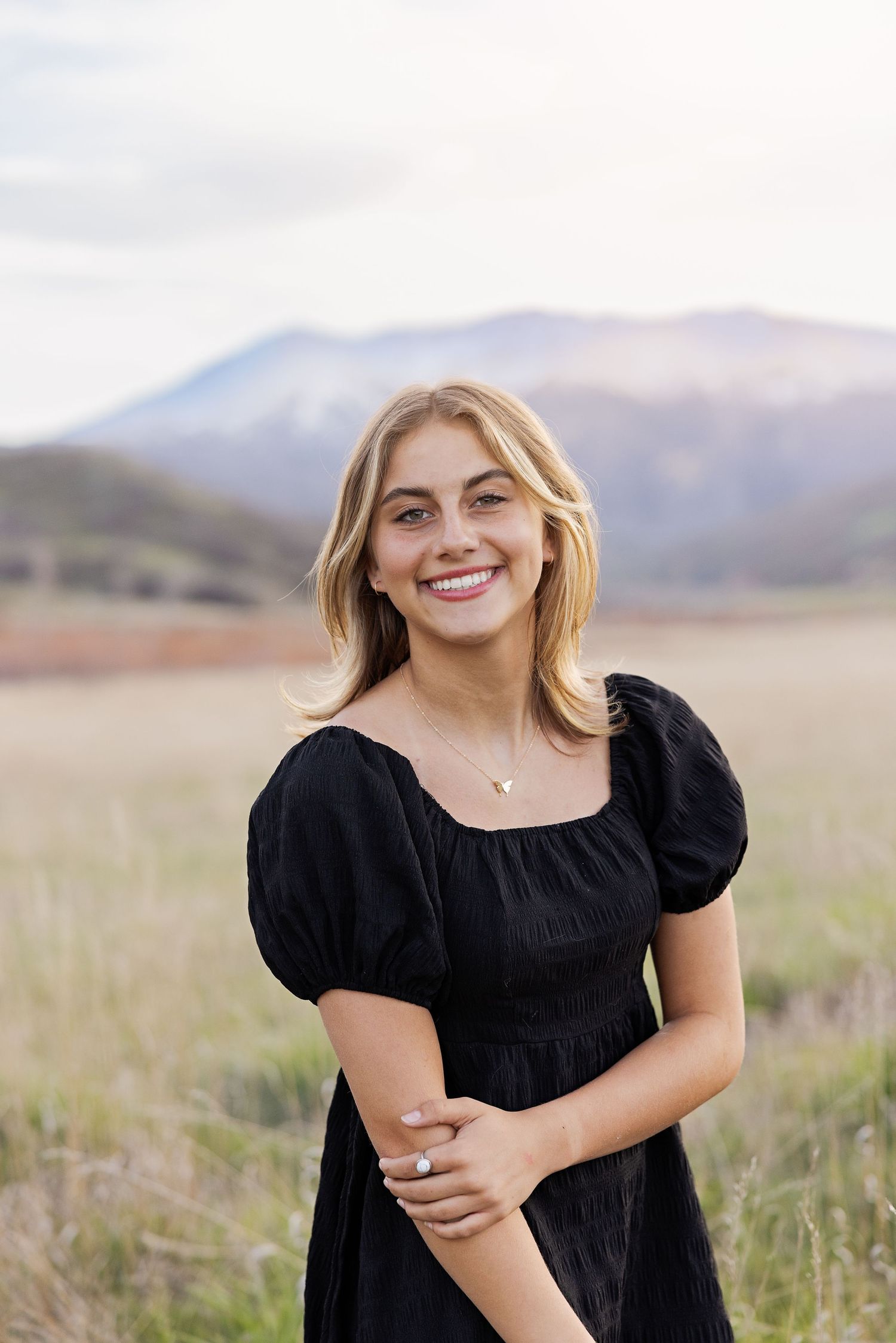 Young blonde woman in black dress smiling outdoors with mountain landscape background.