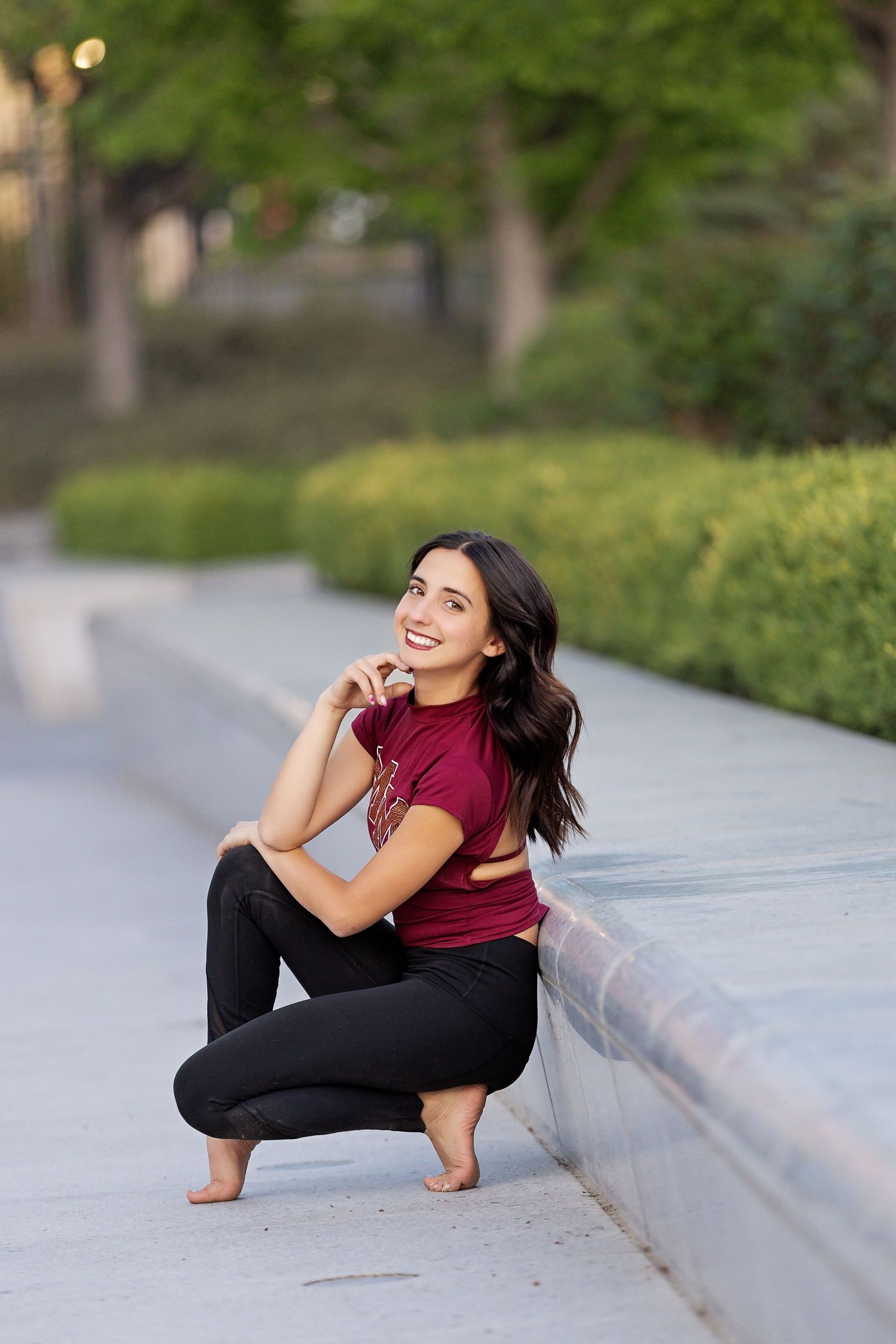Athletic dancer in burgundy top and black pants performs expressive poses on concrete ledge in park setting.