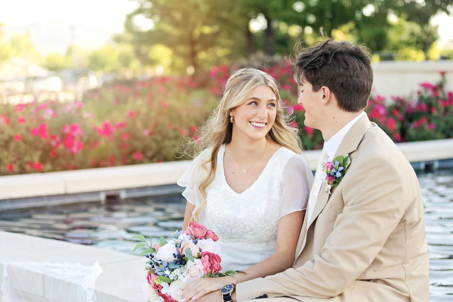 Romantic wedding couple sits by a lakeside garden with pink flowers blooming in the background during sunset.