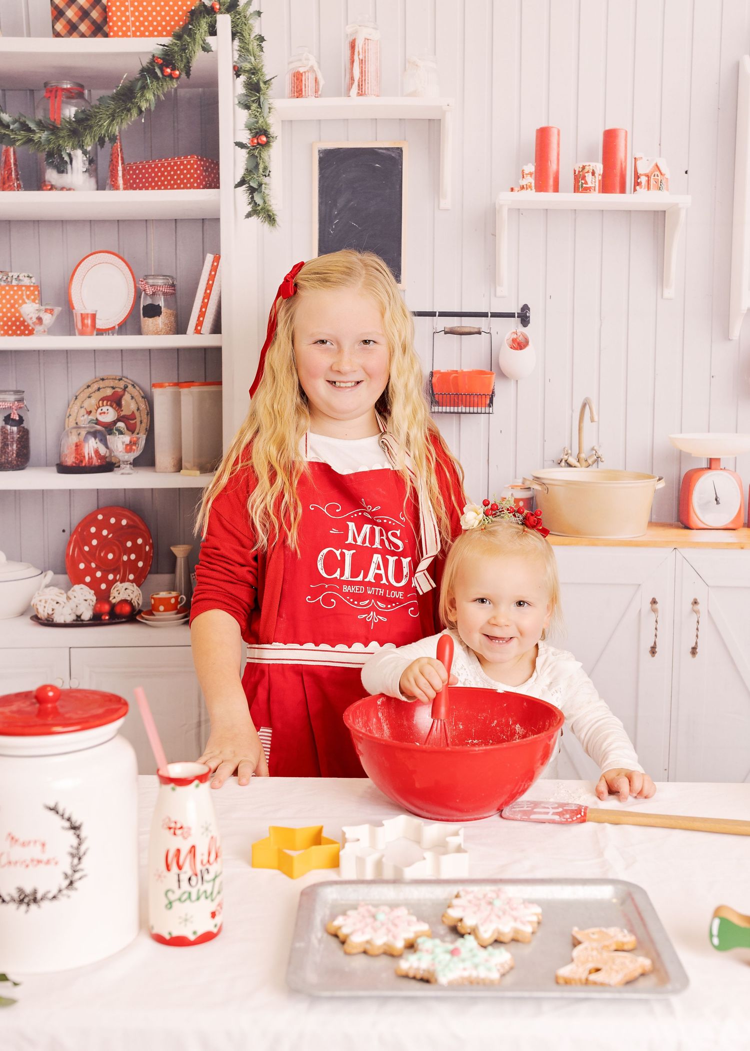 Two children baking Christmas cookies in a festive red and white kitchen with holiday decorations and baking supplies.