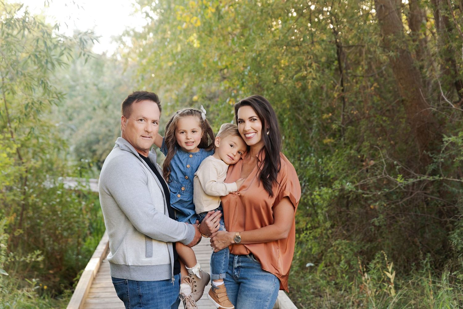 A family of four poses together on a wooden boardwalk surrounded by greenery in autumn.