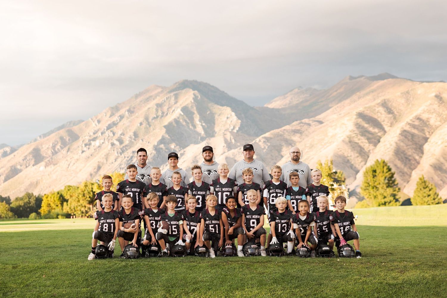 High school football team poses for panoramic photo on grass field with scenic mountain backdrop at sunset.