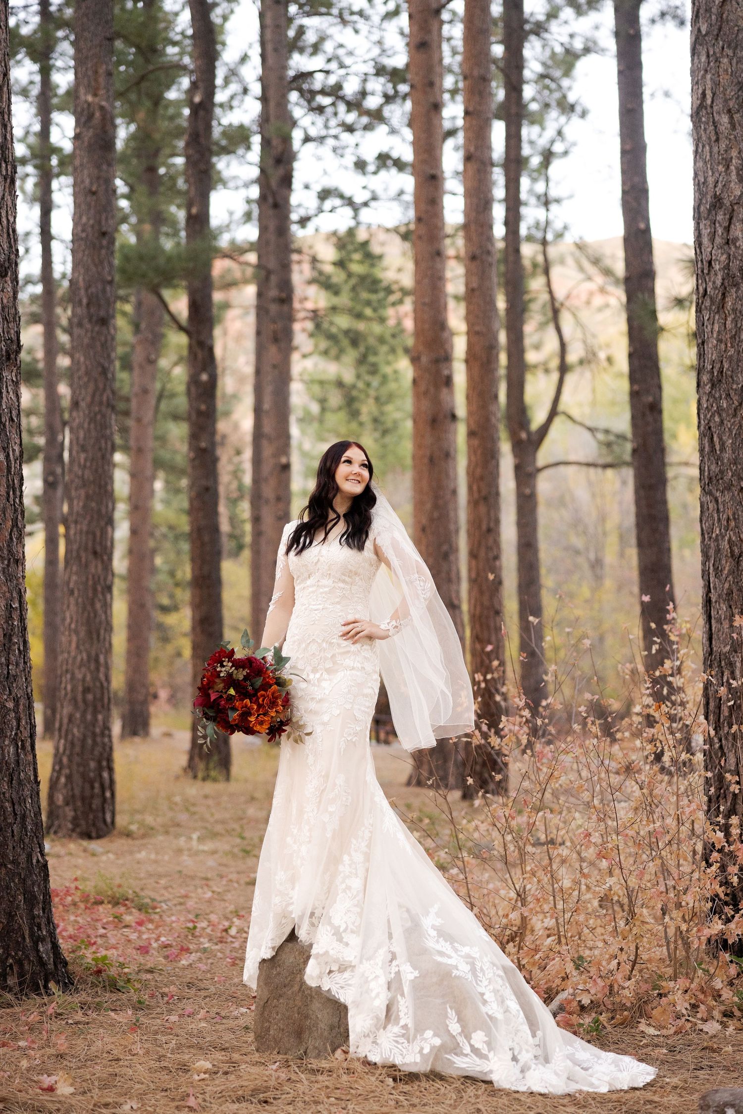 Stunning bridal portrait among tall pine trees with flowing lace gown and vibrant fall bouquet.