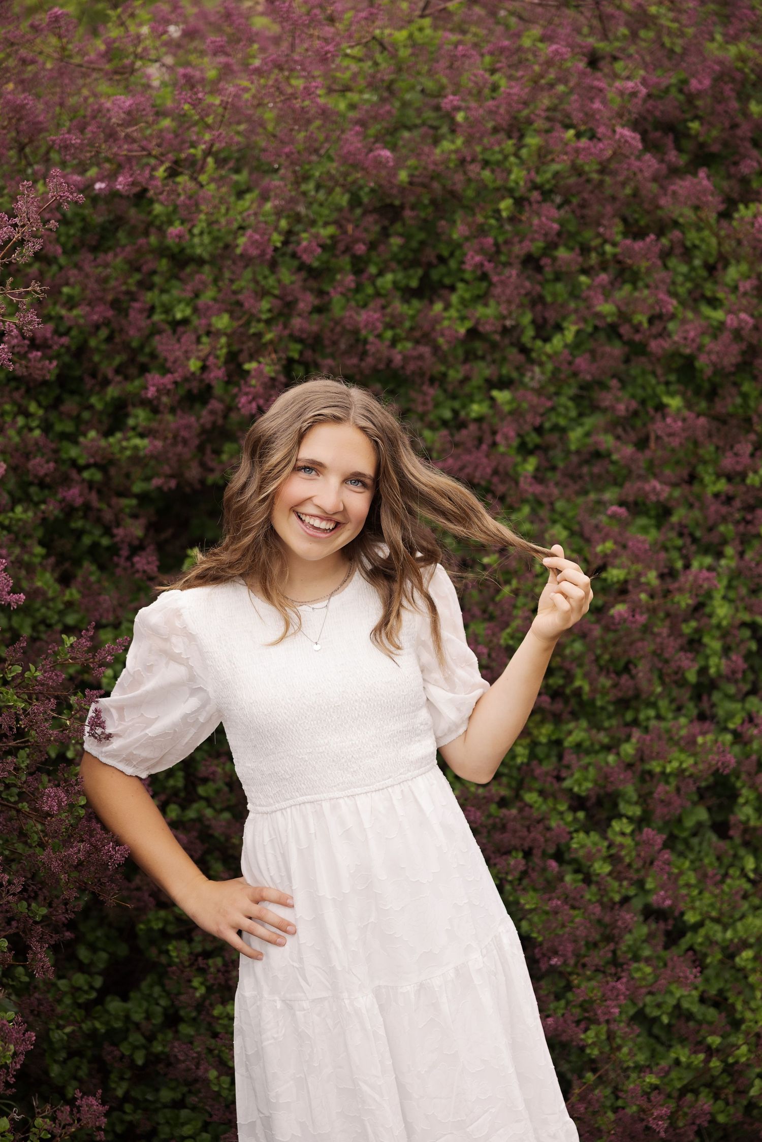 Woman in white dress poses playfully against blooming purple flowers.