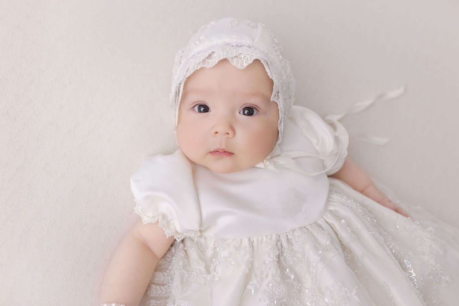 Baby dressed in white baptism gown and lace bonnet on light background.
