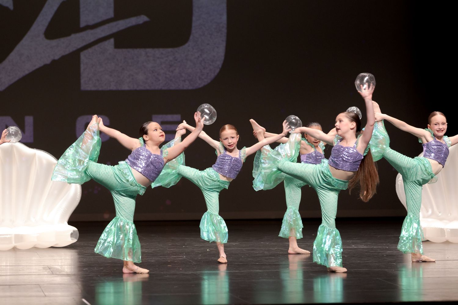 Dancers in flowing mint green costumes perform synchronized leg movements on stage during a dance recital performance.