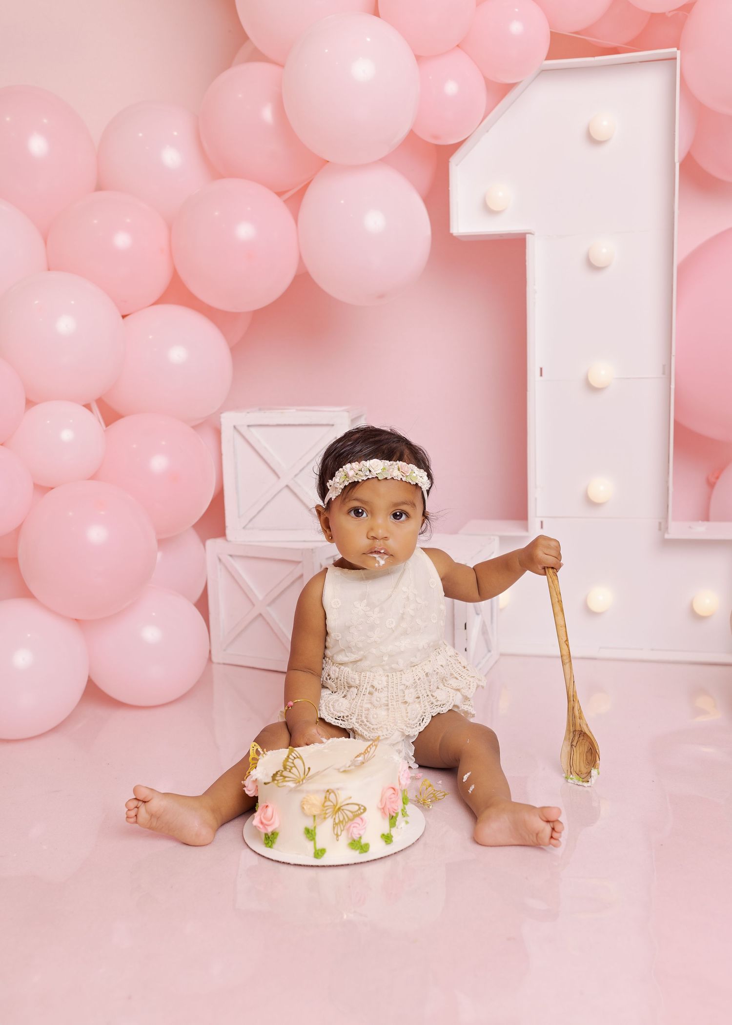 Baby in white dress sits with birthday cake against pink balloon backdrop during first birthday photoshoot.
