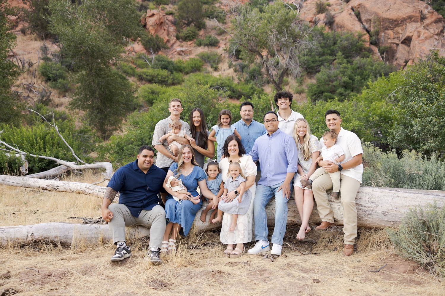 Large family group portrait outdoors in a natural desert landscape with red rocks and greenery in the background.