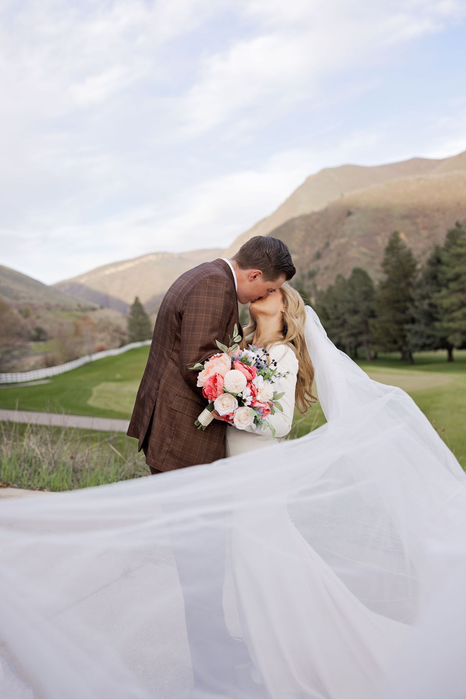 Newlyweds share romantic moment on a mountainside, with flowing white veil and pink floral bouquet in scenic landscape.
