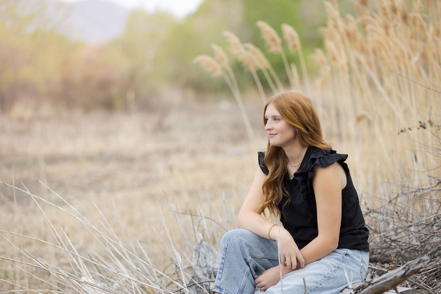 Young woman with red hair sitting in a field of tall grasses, wearing a black top and jeans, smiling softly.