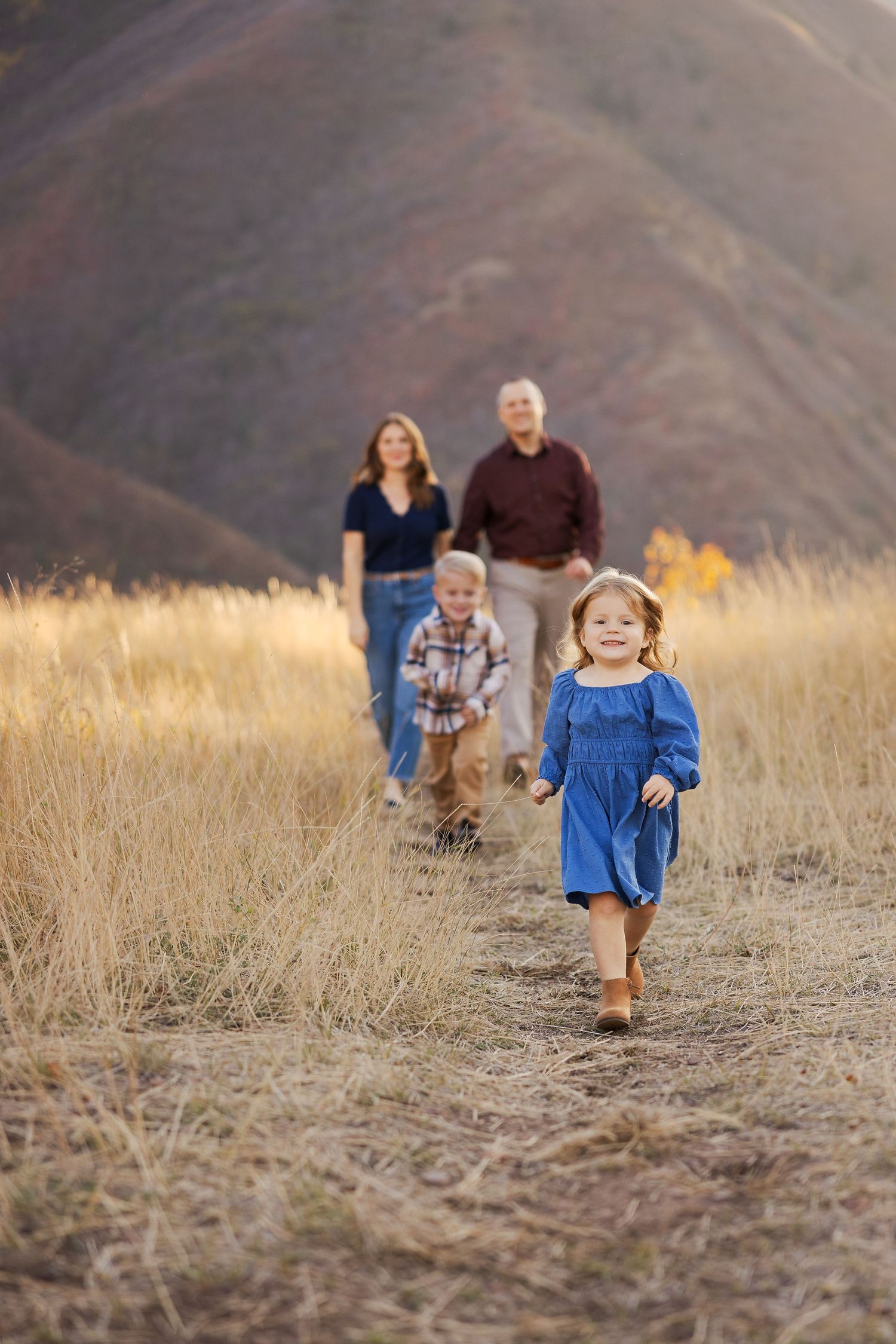 Family walks along mountain trail as children run ahead through golden grass at sunset.