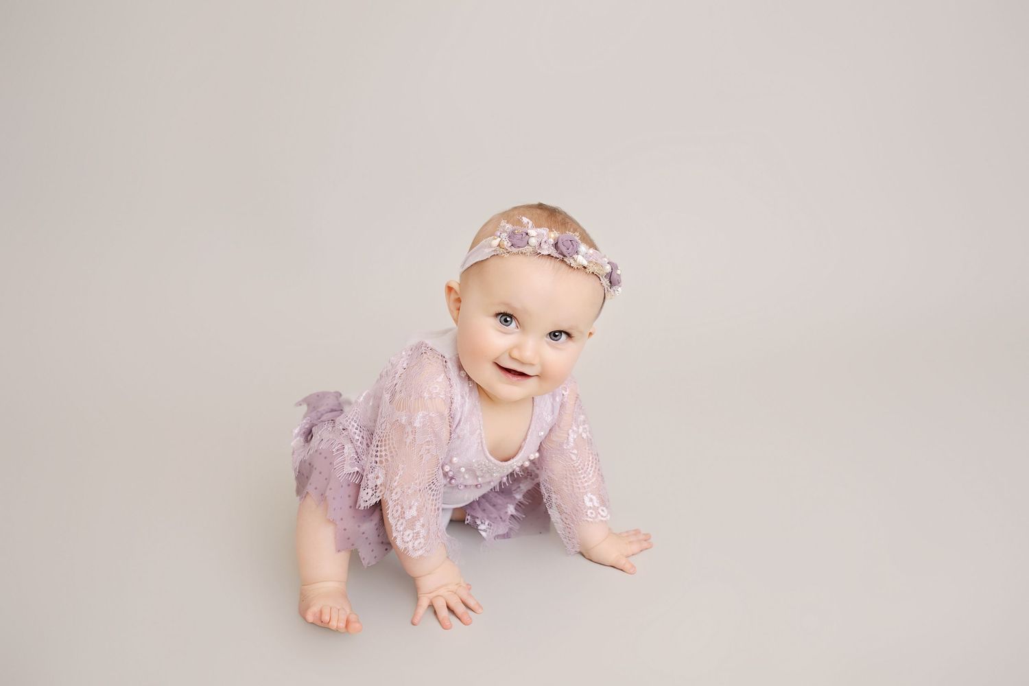 A baby in a purple outfit crawling on a neutral studio backdrop.