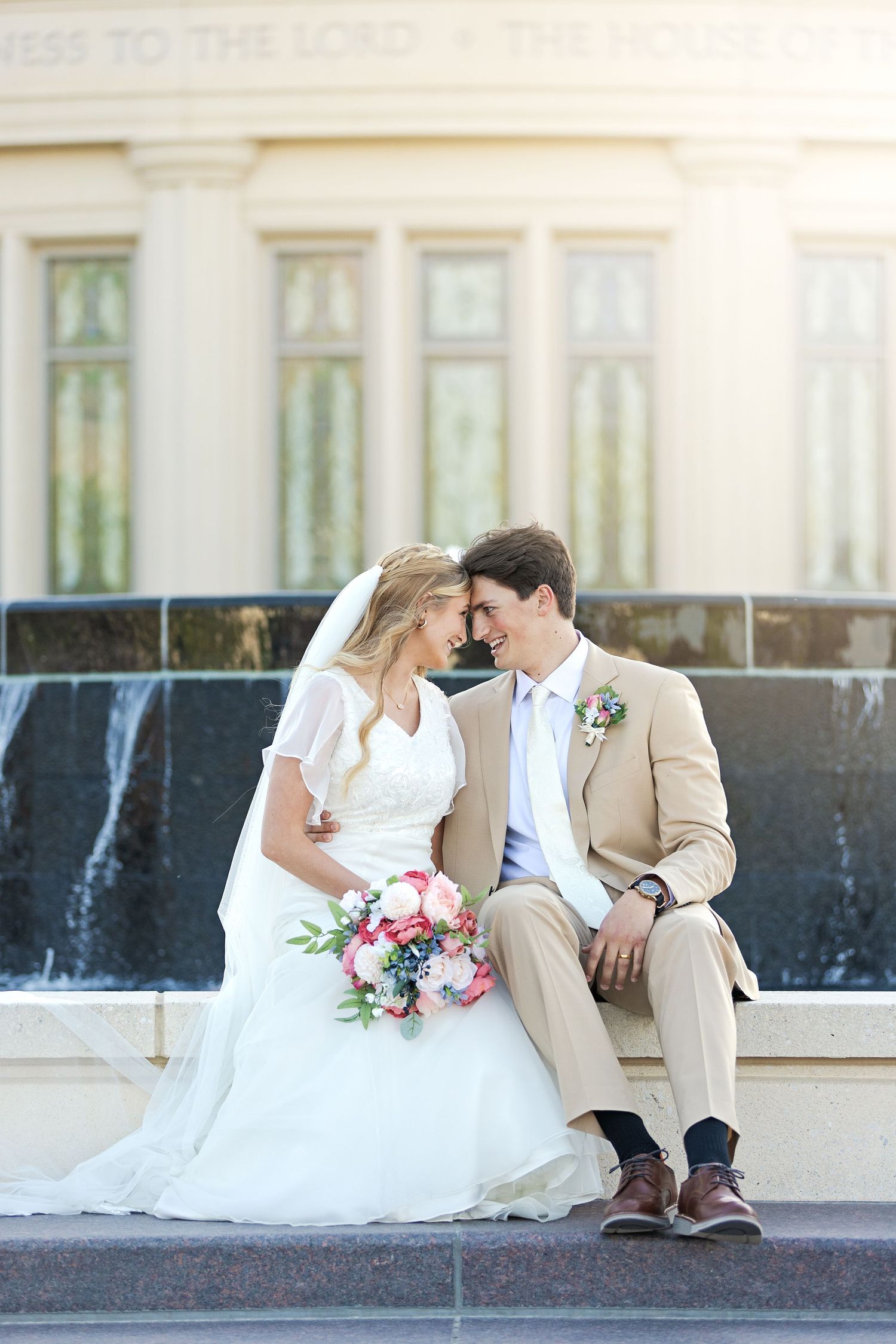 A wedding couple sits by an outdoor fountain in romantic poses wearing white and beige attire with pink floral bouquet.