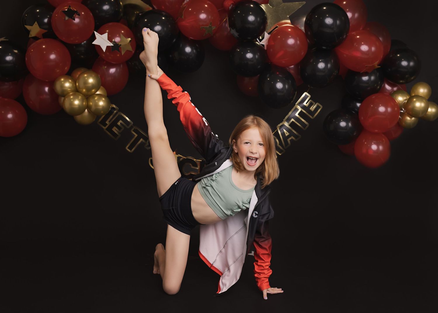 Dynamic dance pose against black backdrop with red and gold balloon arch decorations.