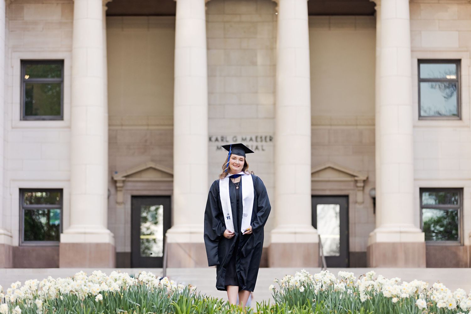 Graduate in black cap and gown stands before a classical stone building with columns and blooming white flowers.