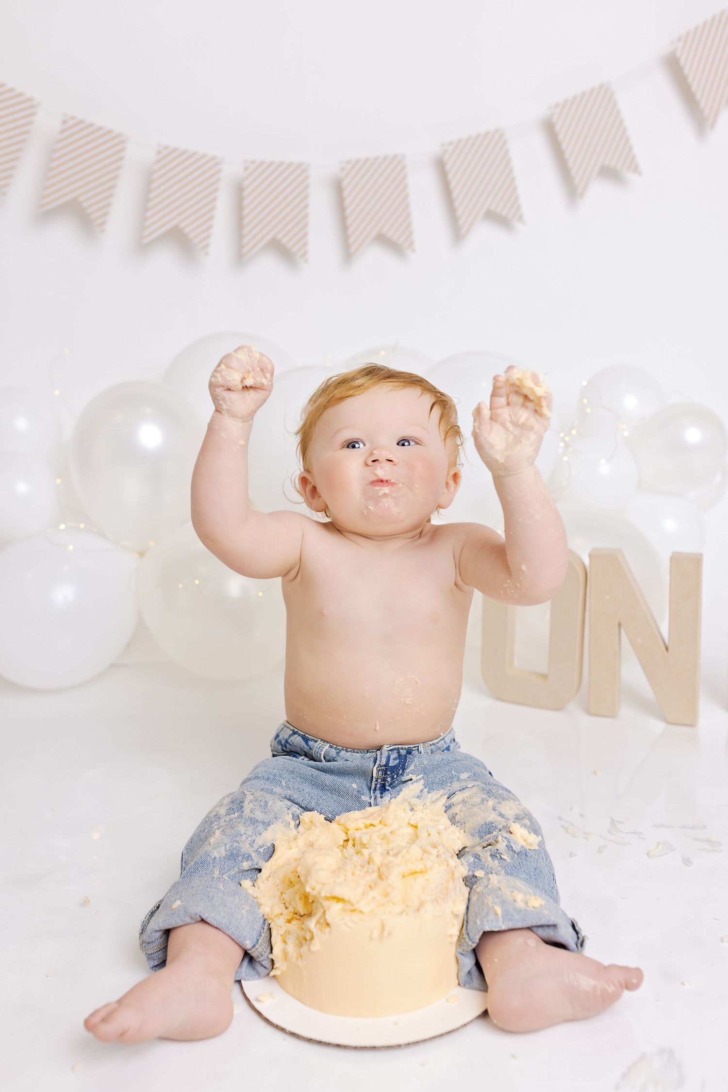 Baby in denim shorts sits with arms raised beside smashed yellow birthday cake against white backdrop with bunting.