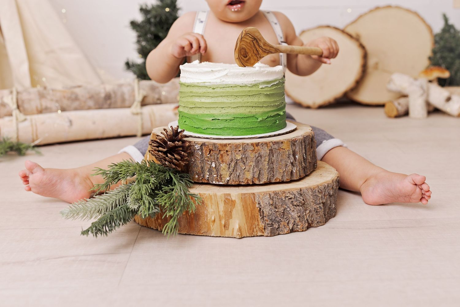 Small child smashing a green and white buttercream cake displayed on rustic wood slices with pine cone decor.