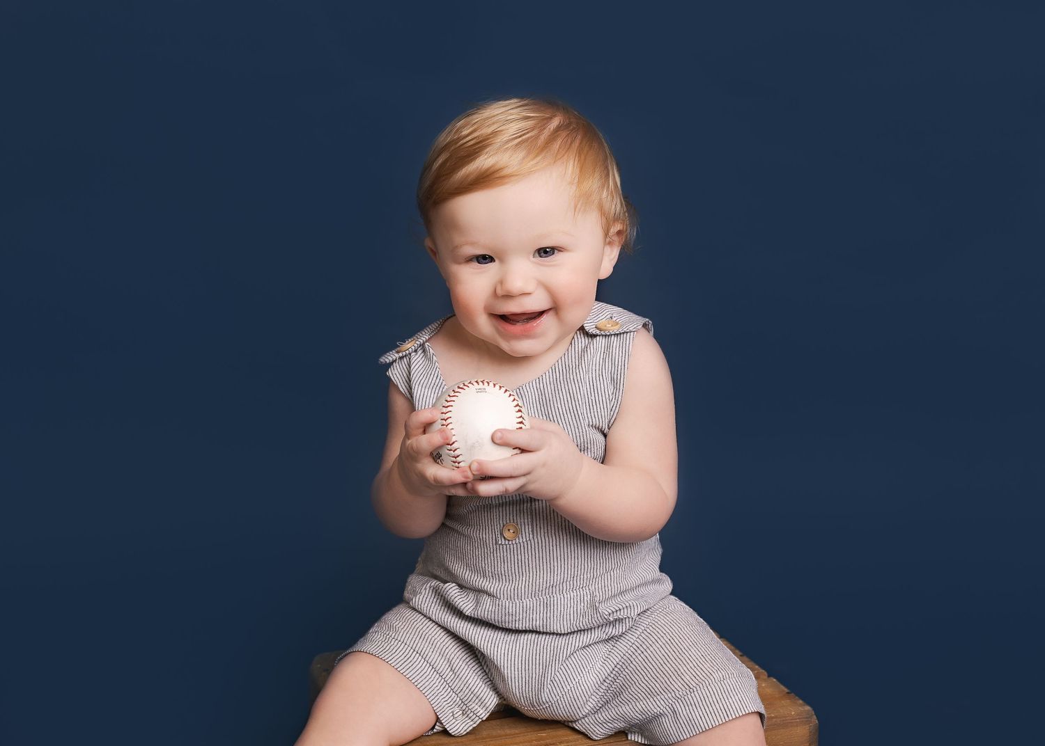 Baby in gray romper smiles while holding a baseball against navy background.