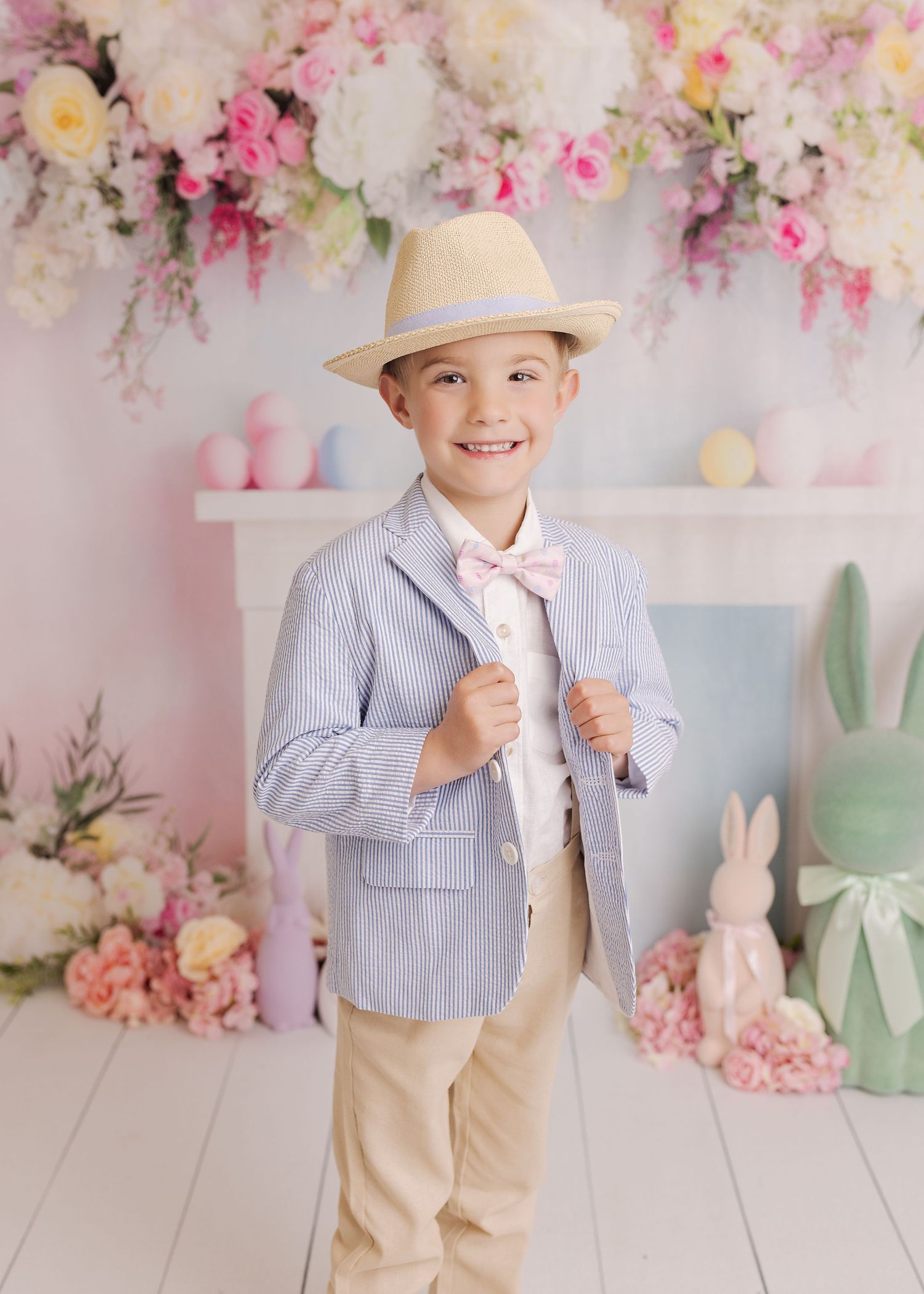 A young child in seersucker blazer and khaki pants poses against a floral backdrop with spring decorations.
