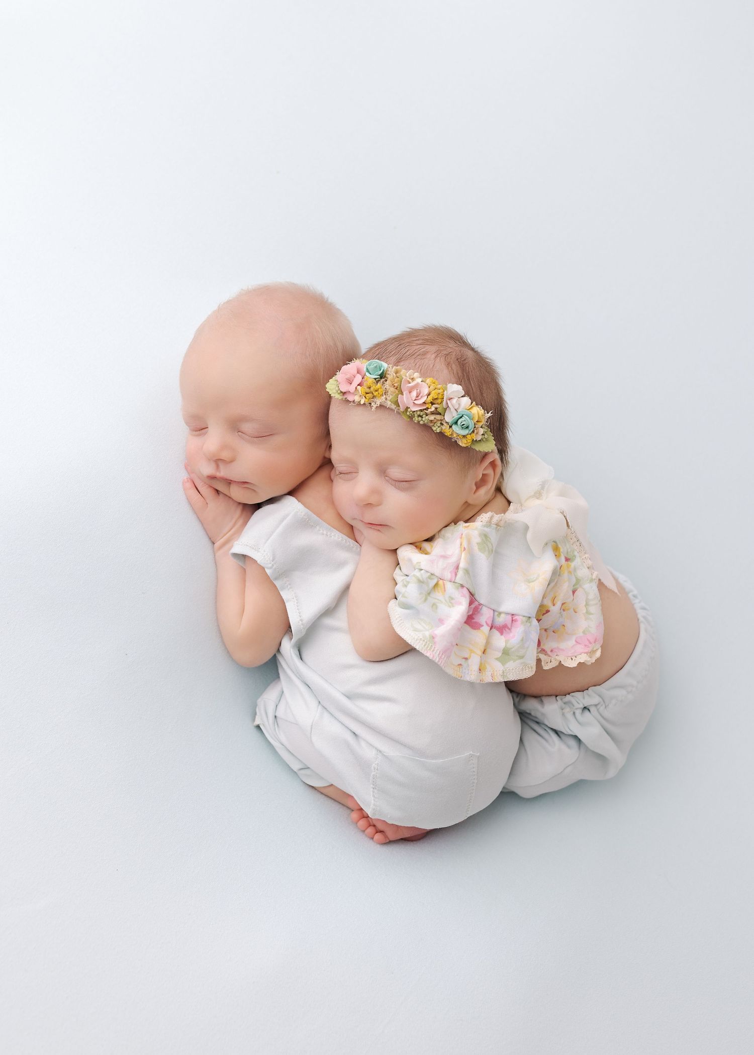 Two sleeping newborn babies posed together wearing white outfits, with one baby wearing a floral headband.