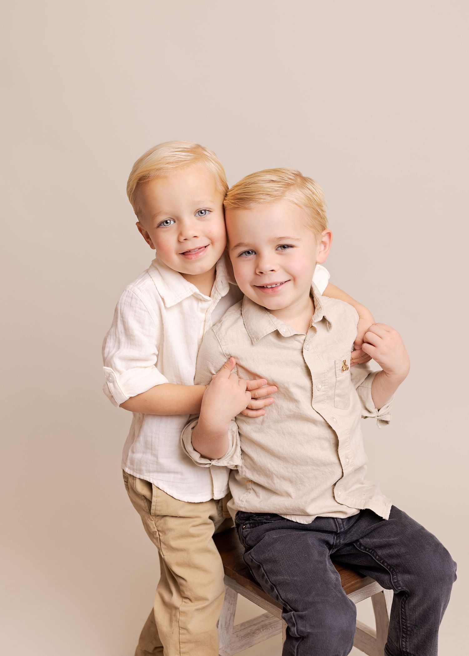 Two children in light colored clothing sitting and playing together on a beige studio backdrop.