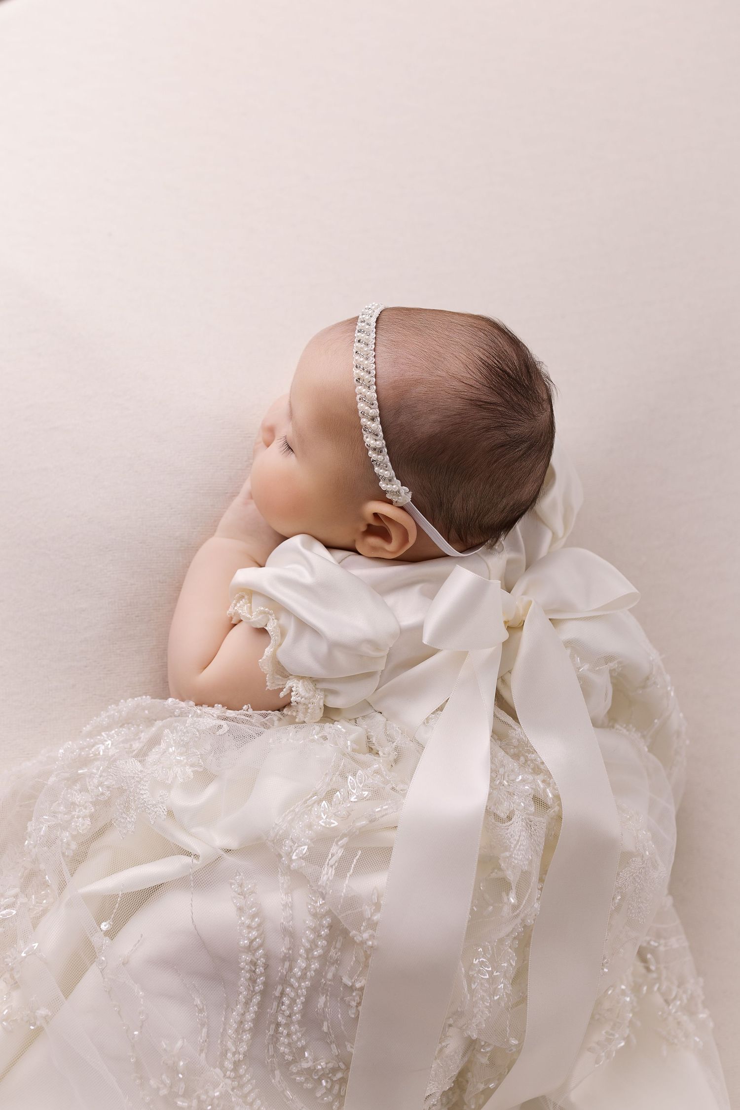 A baby in a delicate white christening gown with lace details and satin bow lies against a light backdrop.