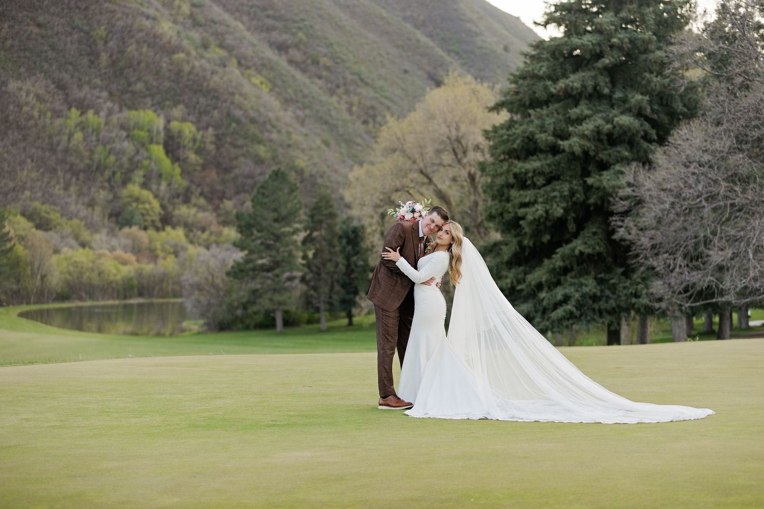 A romantic wedding moment captured on a golf course with mountains and evergreen trees in the background.