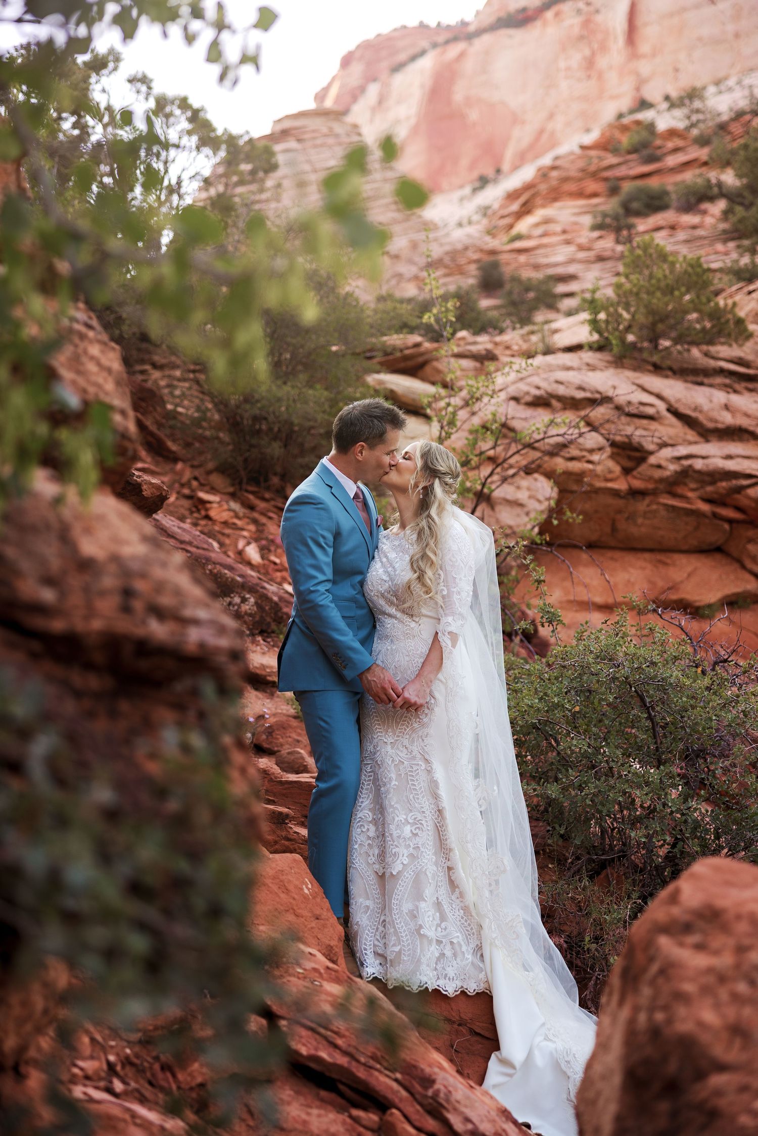 A romantic wedding portrait of a couple on red rock cliffs surrounded by desert vegetation.